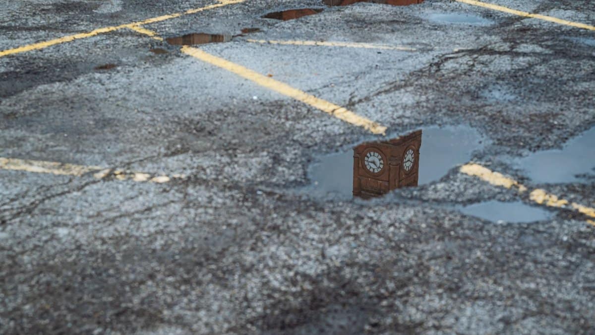 A clock tower reflects in puddles on a wet asphalt road, showcasing urban textures.