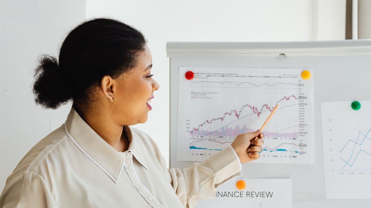A woman points at a stock market chart on a whiteboard, explaining financial trends.