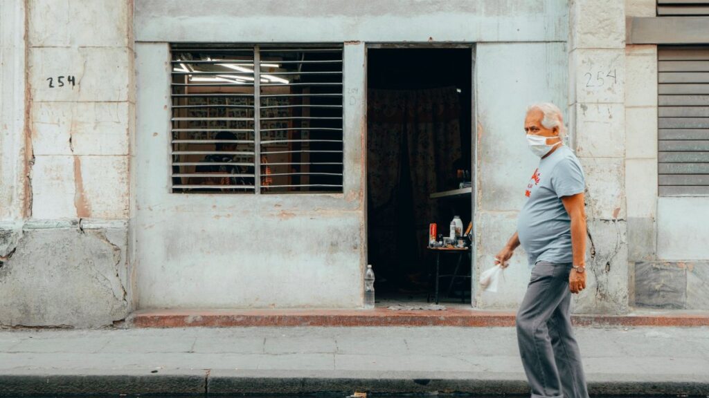 Senior man wearing mask walking past a weathered building in Havana, Cuba, during daytime.