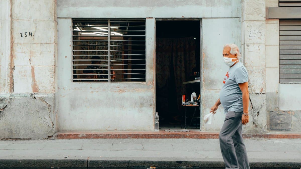 Senior man wearing mask walking past a weathered building in Havana, Cuba, during daytime.
