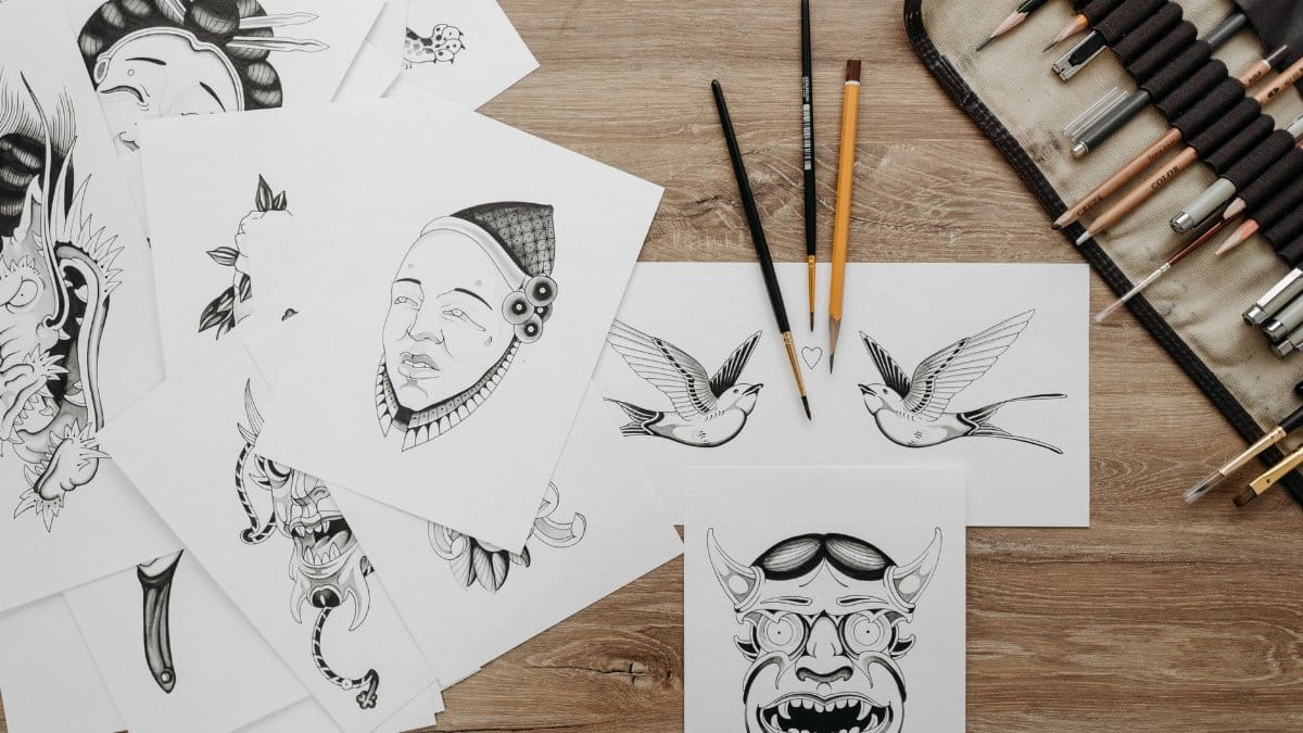 Flat lay of intricate tattoo sketches and drawing tools on wooden table.