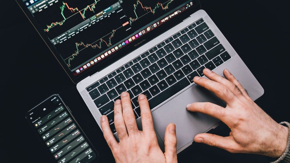 Hands typing on a laptop displaying stock market charts and a smartphone with financial apps.