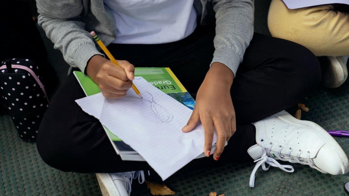 From above of crop ethnic schoolkid drawing with pencil on paper sheet while sitting with crossed legs on pavement near anonymous friend