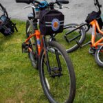 Group of orange bicycles resting on green grass by a paved trail in an outdoor park setting.