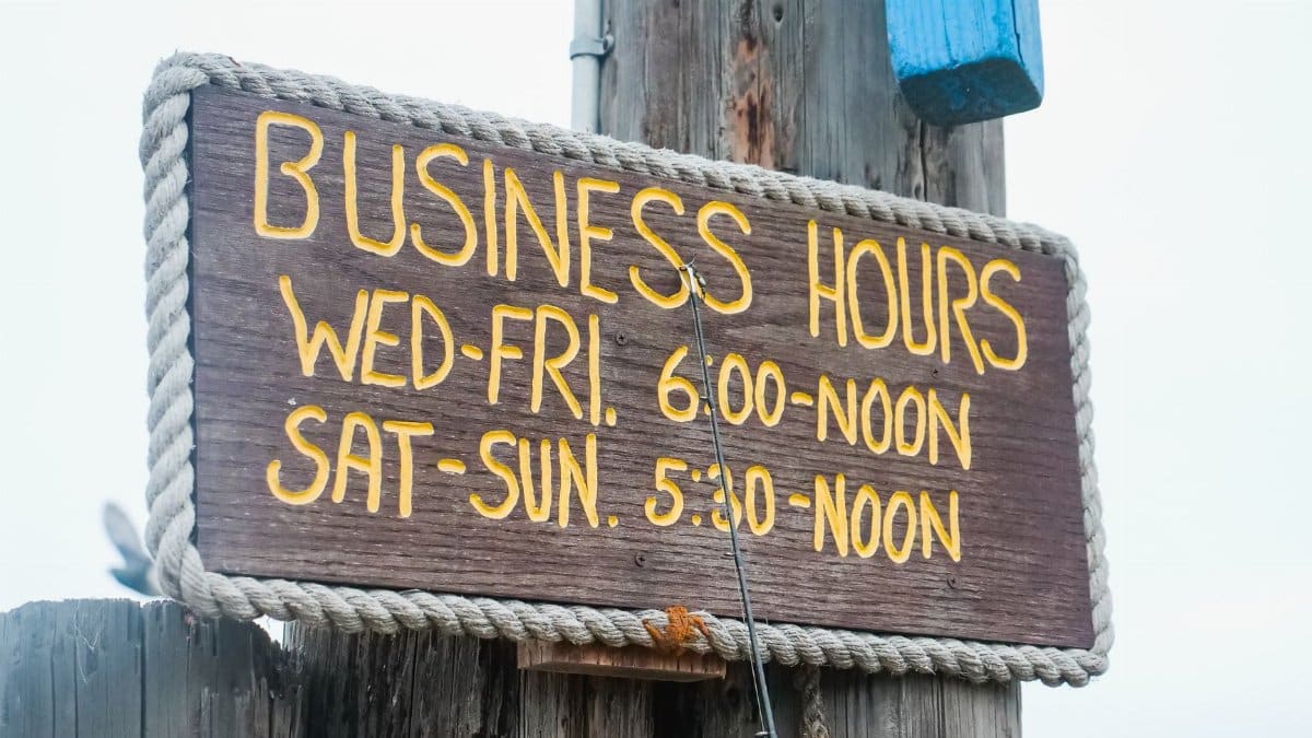 Weathered wooden sign displaying business hours with bright yellow text outdoors.