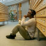 A thoughtful man listens to music sitting in a library aisle, surrounded by vintage card catalogs.
