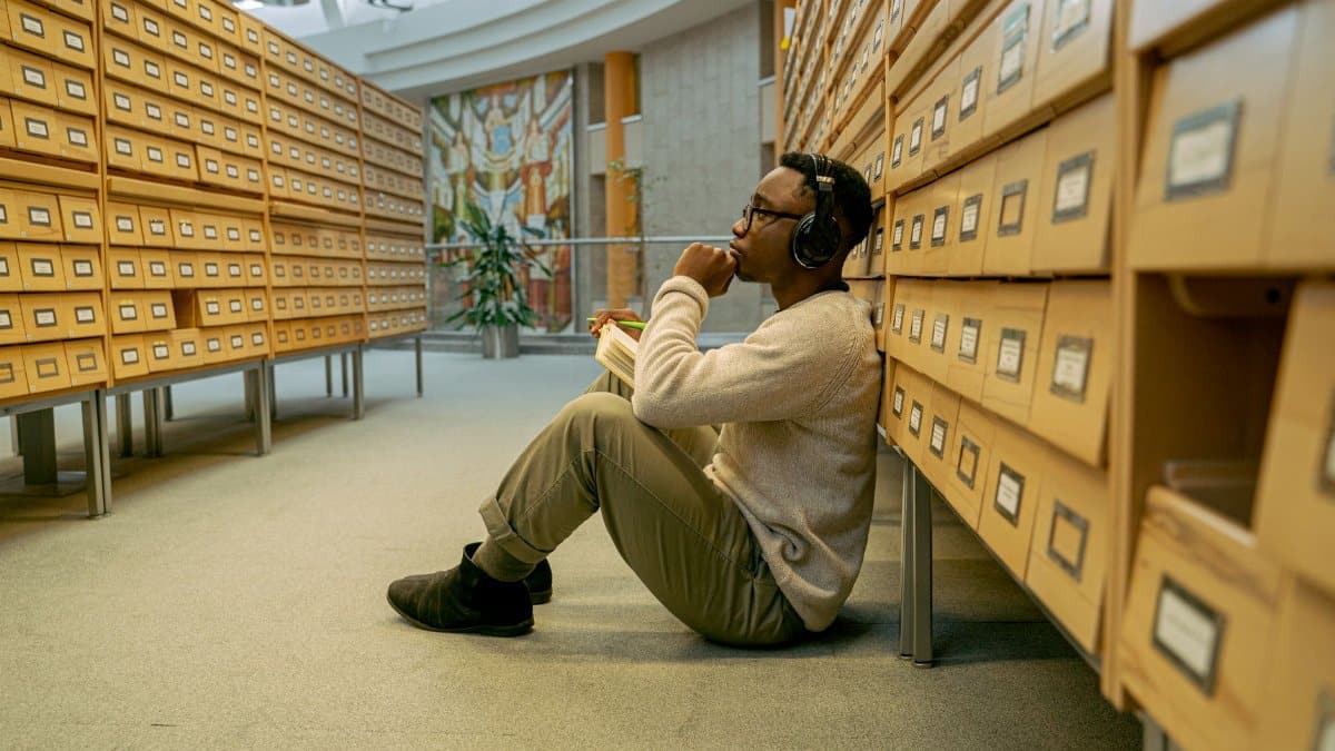 A thoughtful man listens to music sitting in a library aisle, surrounded by vintage card catalogs.