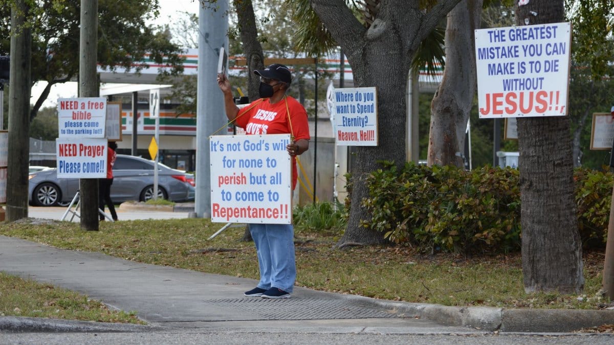 Protester on sidewalk holding signs with religious messages urging repentance.