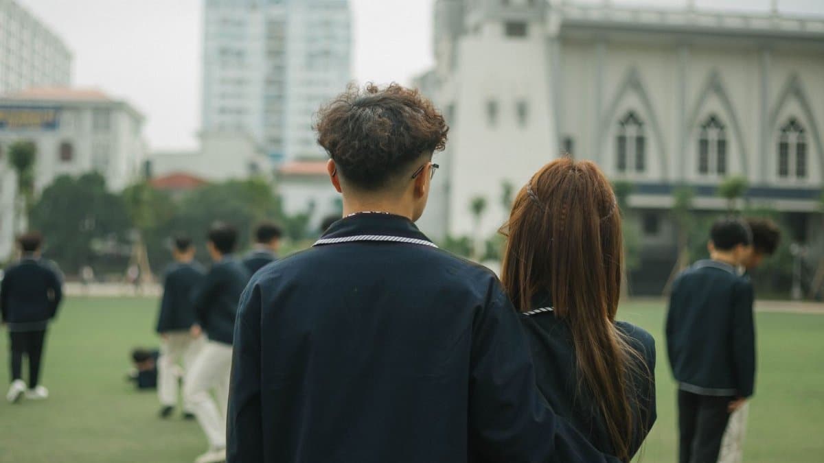 Teen couple in school uniforms embracing on a campus field with peers.