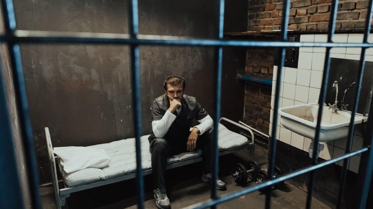 A solitary prisoner in a dimly lit cell, sitting on a bed in deep thought, evoking a sense of isolation and introspection.