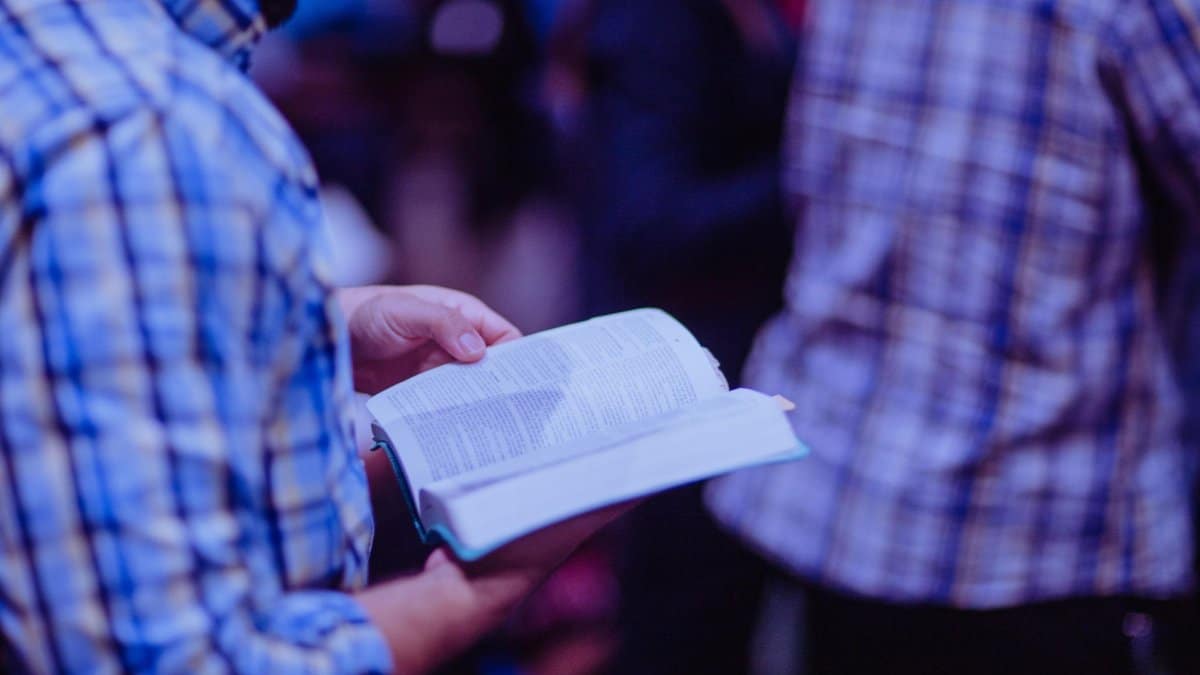 A man stands indoors reading a Bible, focusing amidst a crowd.