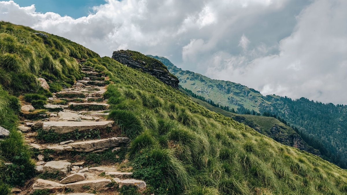 A tranquil hiking path through scenic mountain landscape with lush green hills and cloudy sky.