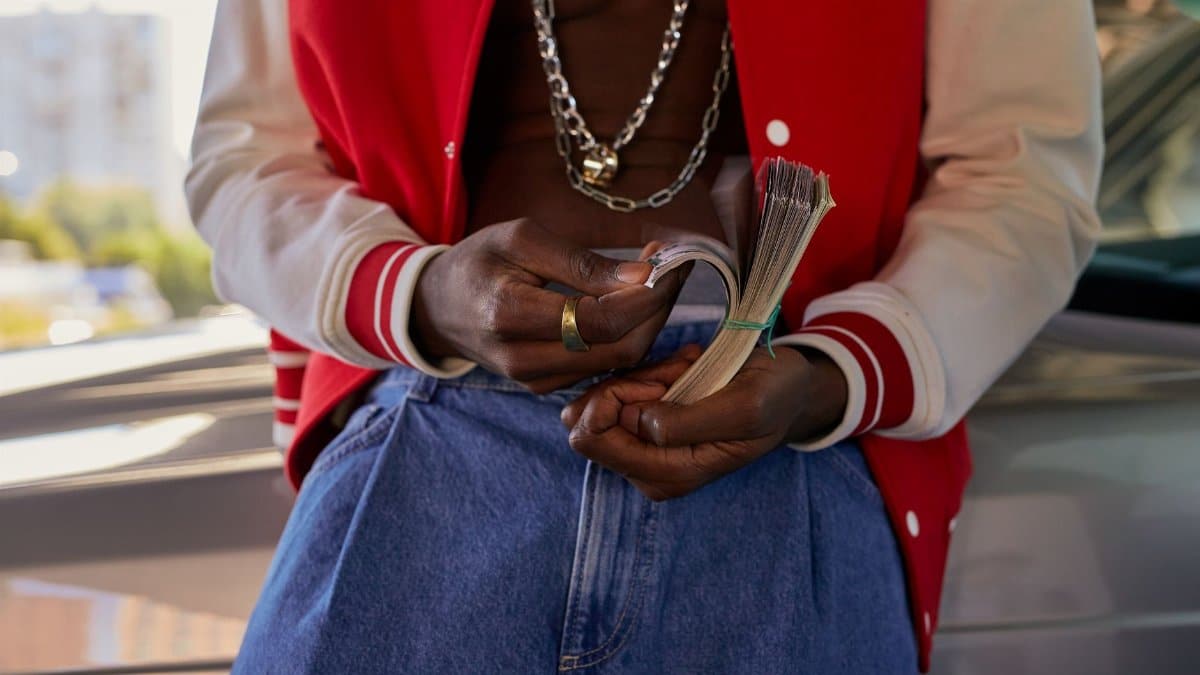 Person in a red jacket holding a stack of cash, wearing jewelry and standing by a car.