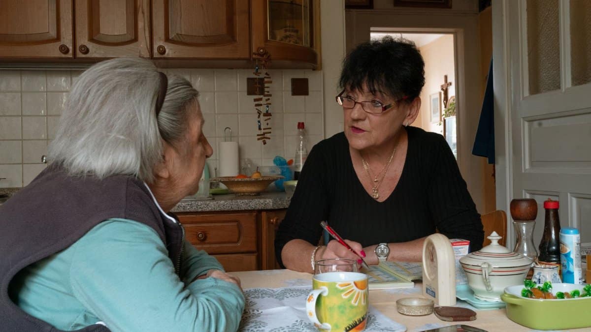 Senior woman and caregiver having a conversation in a cozy Prague kitchen setting.