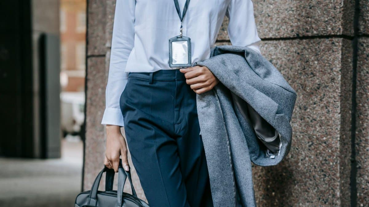 Crop faceless female entrepreneur in stylish formal outfit and name tag on neck walking on city street with briefcase and coat in hands