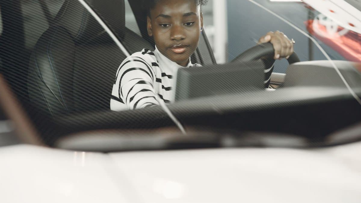 Young woman with black hair driving a car, captured from the interior with focus on the steering wheel.
