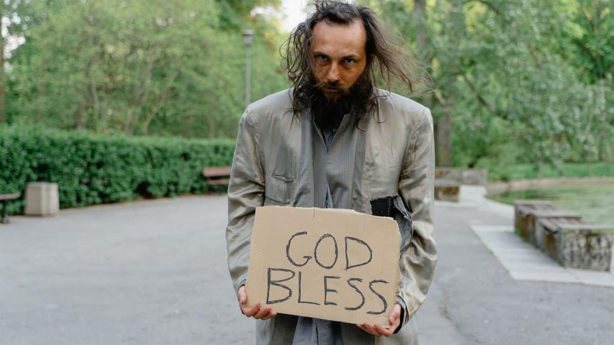 Portrait of a homeless man holding a 'God Bless' sign in a park setting.