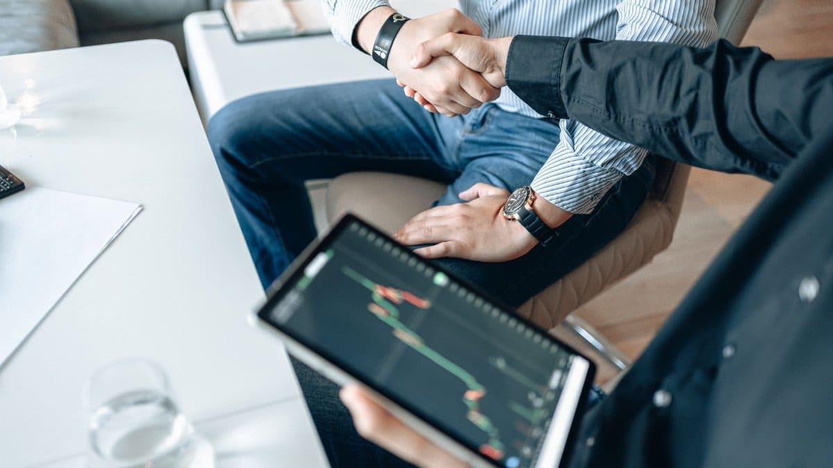 Business professionals shaking hands with a tablet displaying financial graphs.