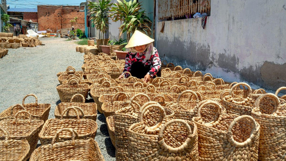 A woman in traditional attire hand-weaving straw baskets on a sunny day outdoors.
