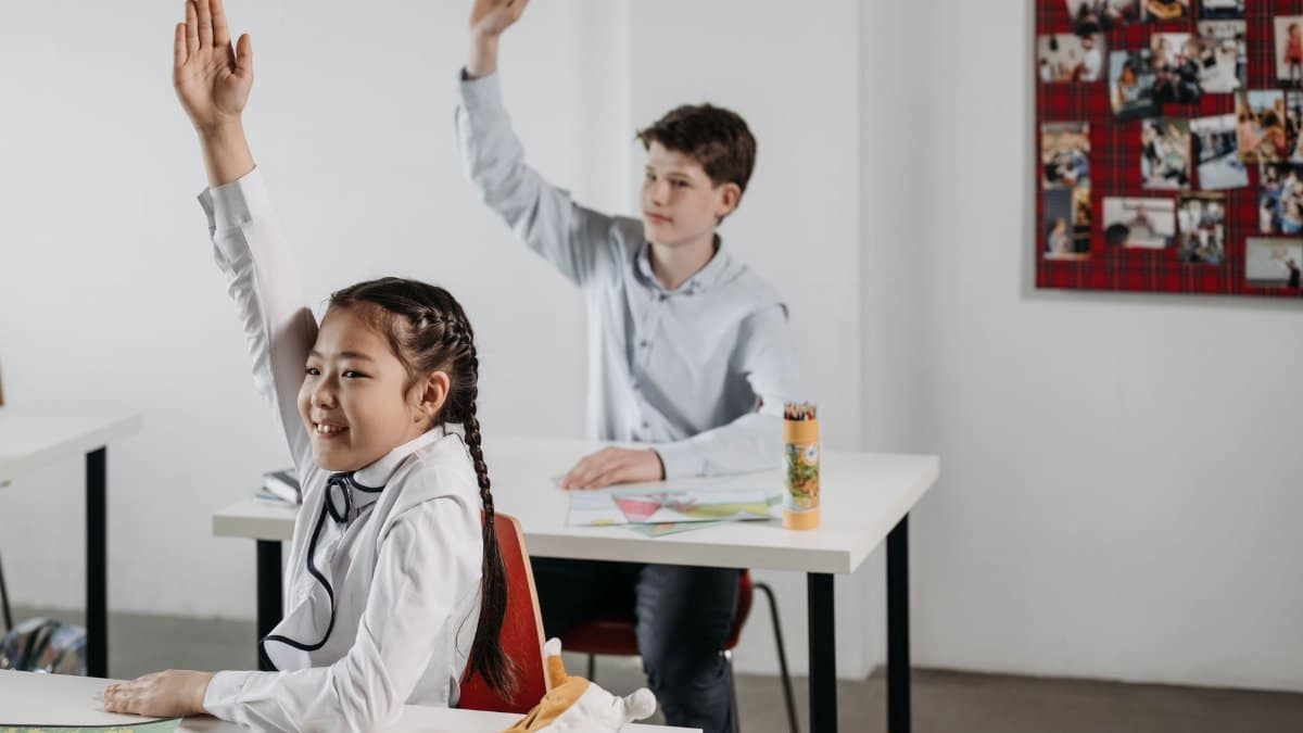 Students actively participating in a classroom setting, seated at desks with raised hands.