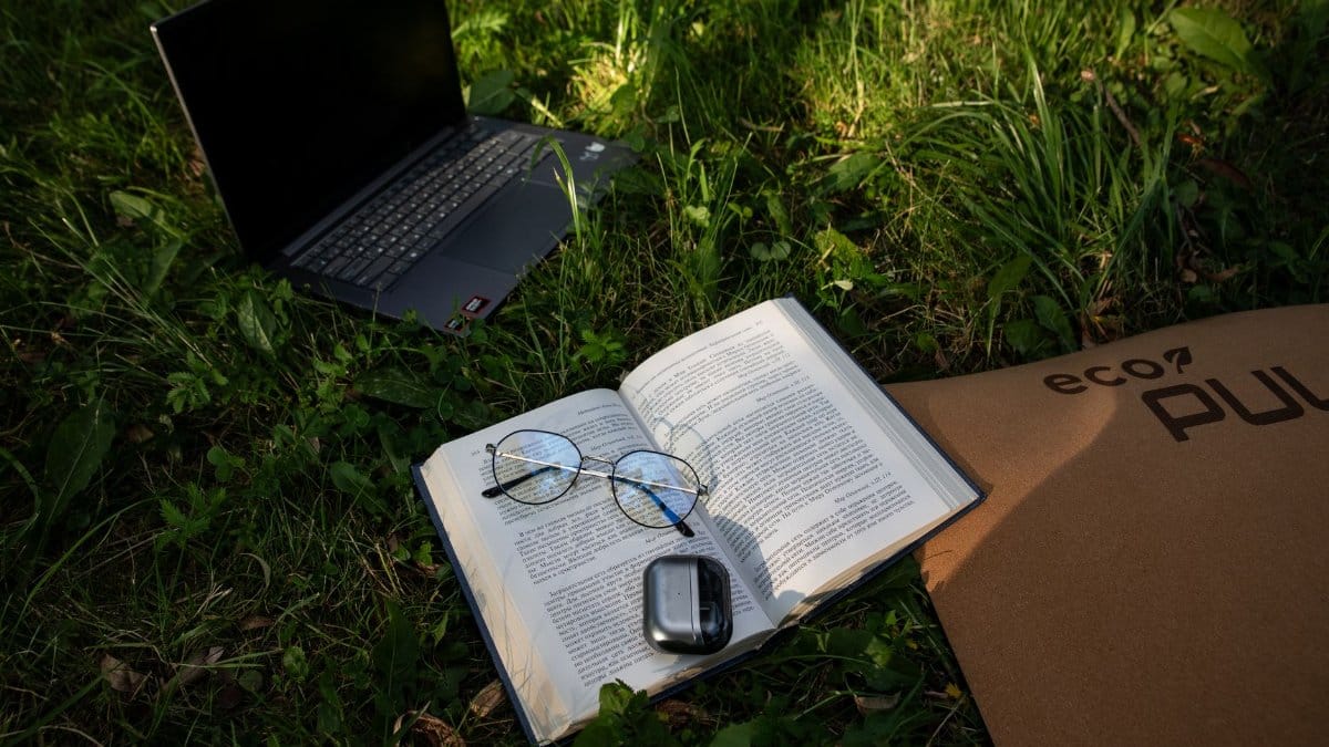 An open book, laptop, and glasses on grass symbolize a serene outdoor work environment.