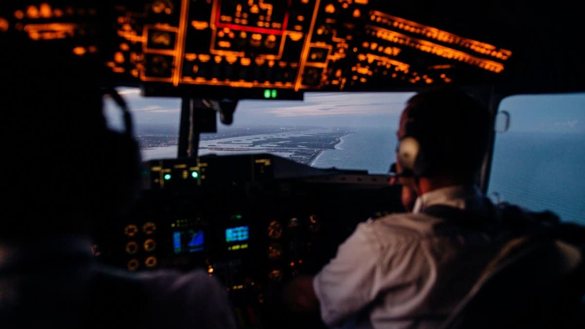 Back view of unrecognizable male pilots in headsets operating illuminating dashboard in cockpit while flying over sea in evening