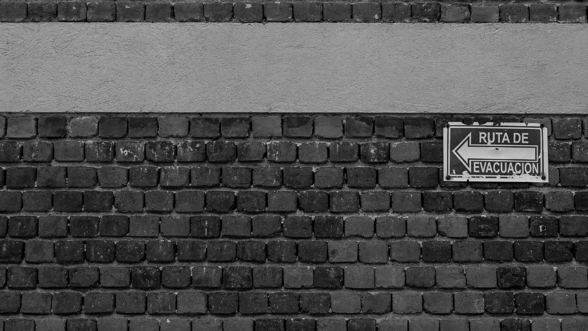 Evacuation route sign on a brick wall in Spanish, captured in black and white.