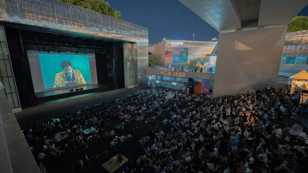 A large audience gathers outdoors at night to watch a movie screening under a bridge.