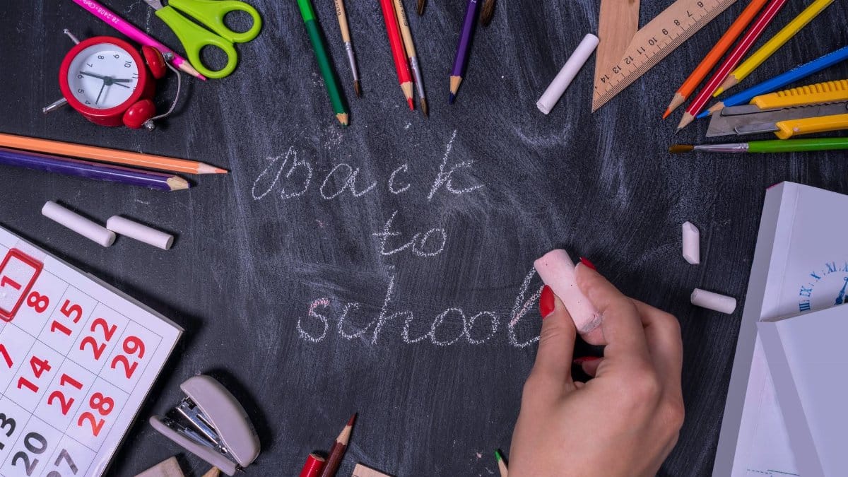 Hand writing 'back to school' on chalkboard surrounded by school supplies.