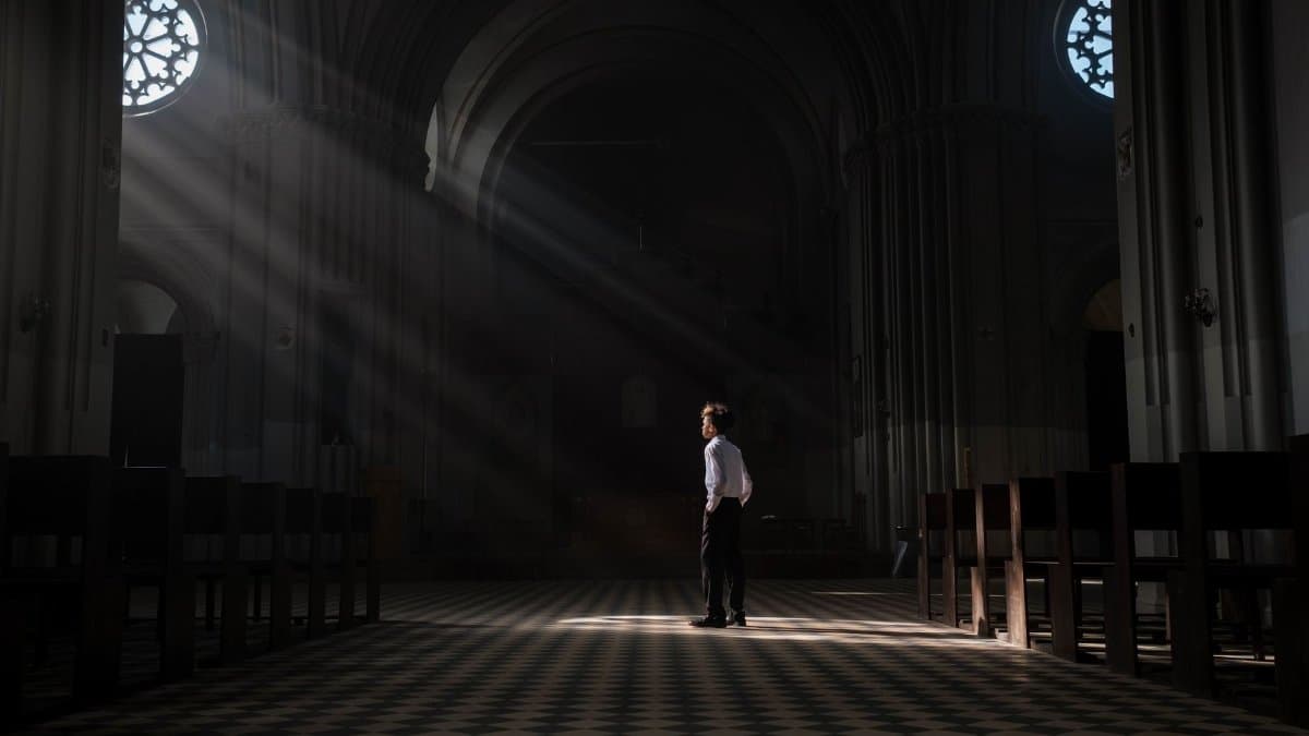 A young boy stands alone in the sunlit interior of a church, casting dramatic shadows.