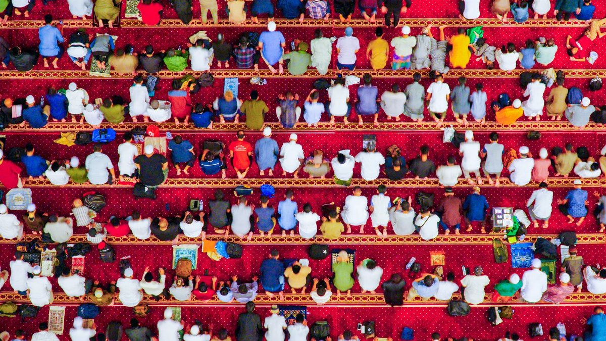 Aerial shot of a congregation sitting on red carpets in a mosque, viewed from above.