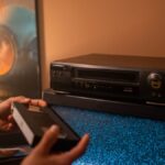 Person inserting VHS tape into video recorder with static TV in cozy room.