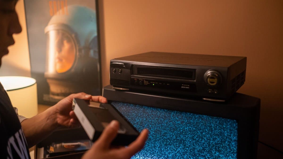 Person inserting VHS tape into video recorder with static TV in cozy room.
