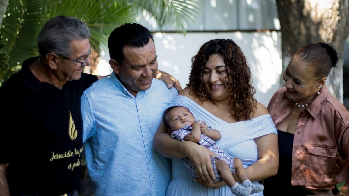 Joyful family gathering with proud parents and grandparents holding a newborn baby outdoors.