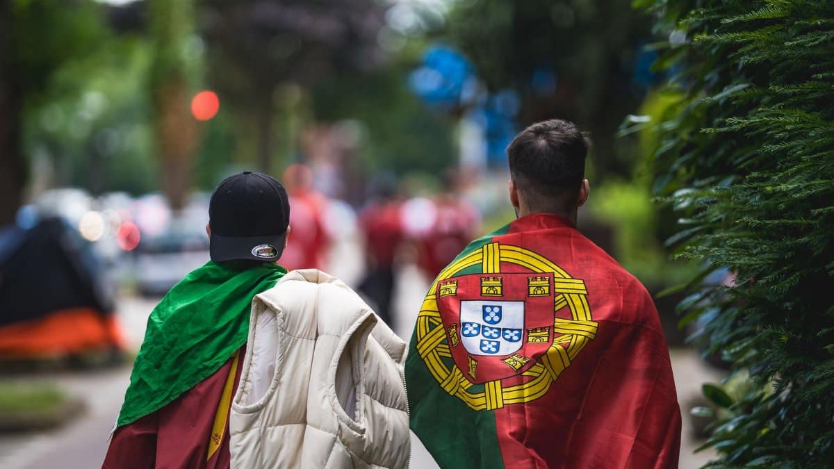 Two sports fans draped in Portuguese flags walking on a city street during daytime.