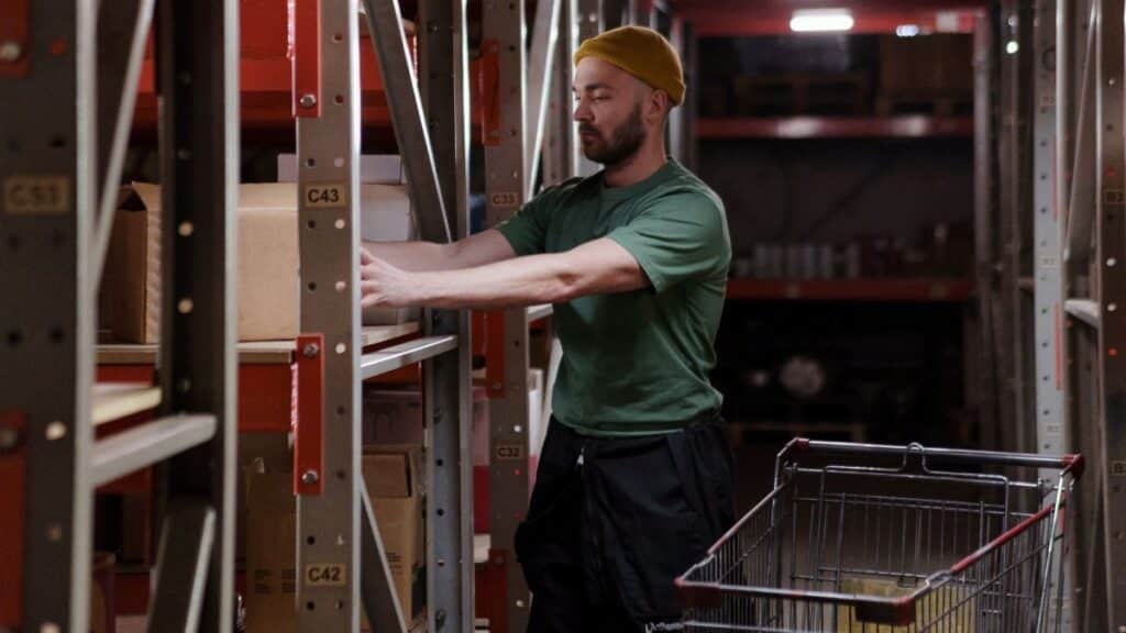 Male warehouse worker in a beanie organizes inventory on shelves with a push cart indoors.