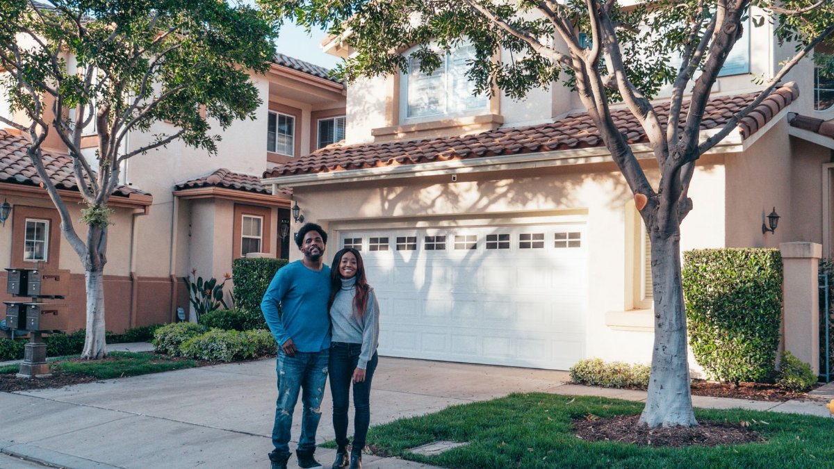 A smiling couple proudly posing in front of their newly purchased house, expressing joy and togetherness.
