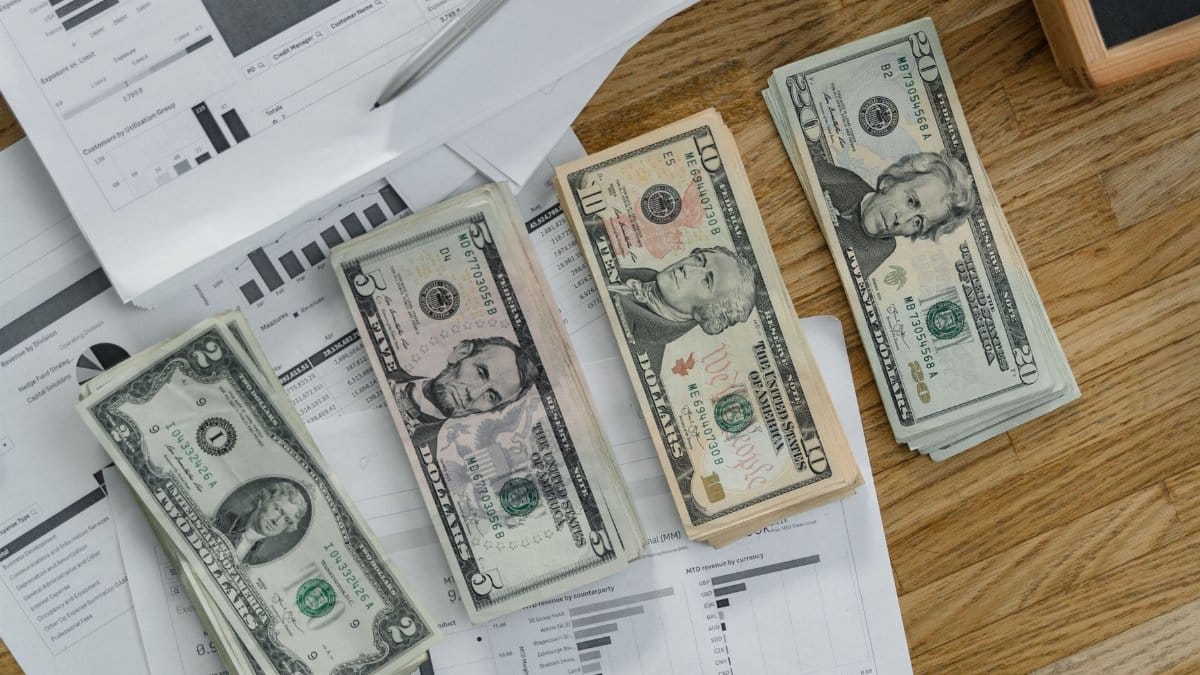 Stacks of US Dollar banknotes placed over financial documents and reports on a desk.