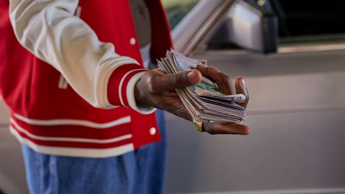 Close-up of a person offering a stack of cash in front of a car, symbolizes financial transaction.