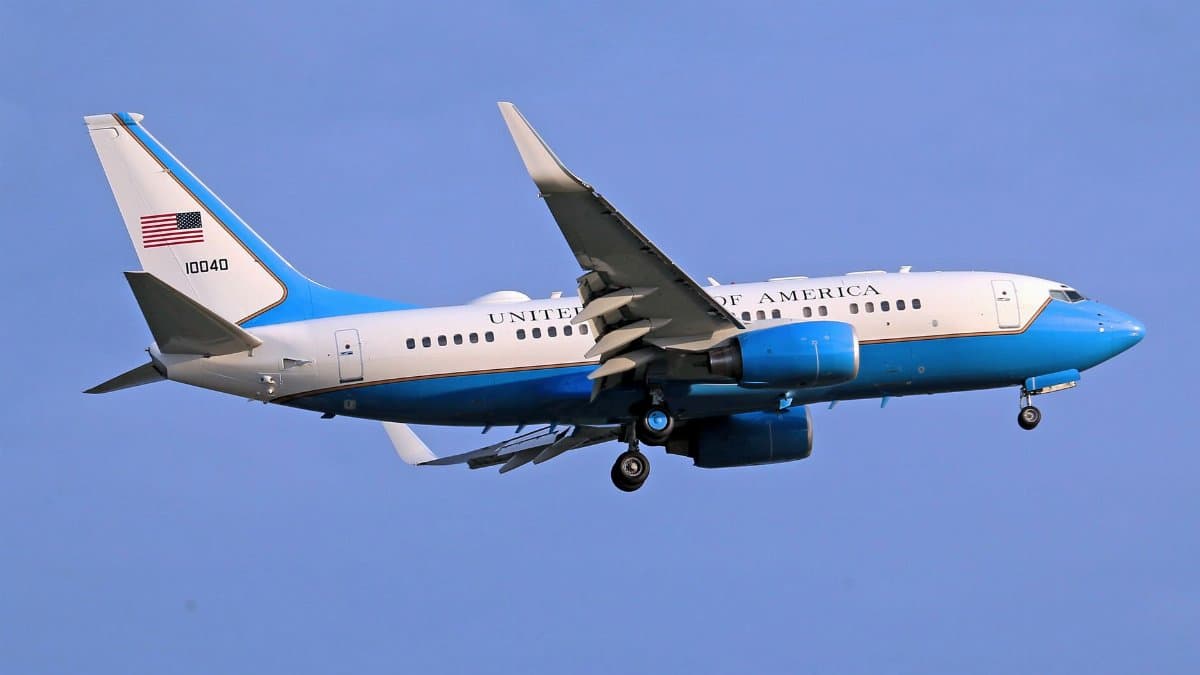 A US government airplane soaring through a clear blue sky, displaying the American flag prominently.