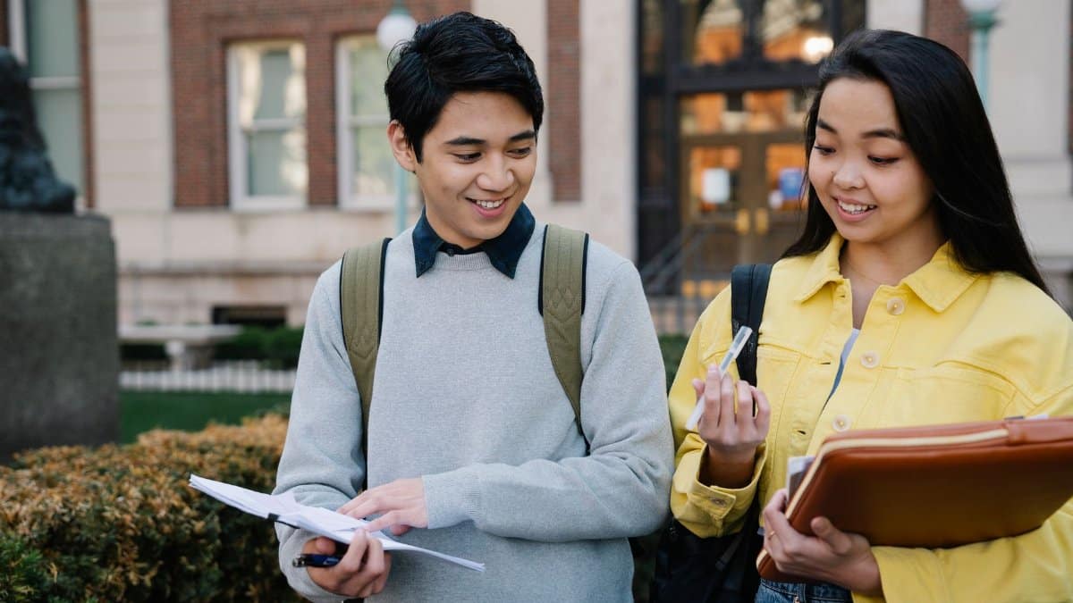 Two students collaborate outdoors on a college campus, sharing smiles and study materials.