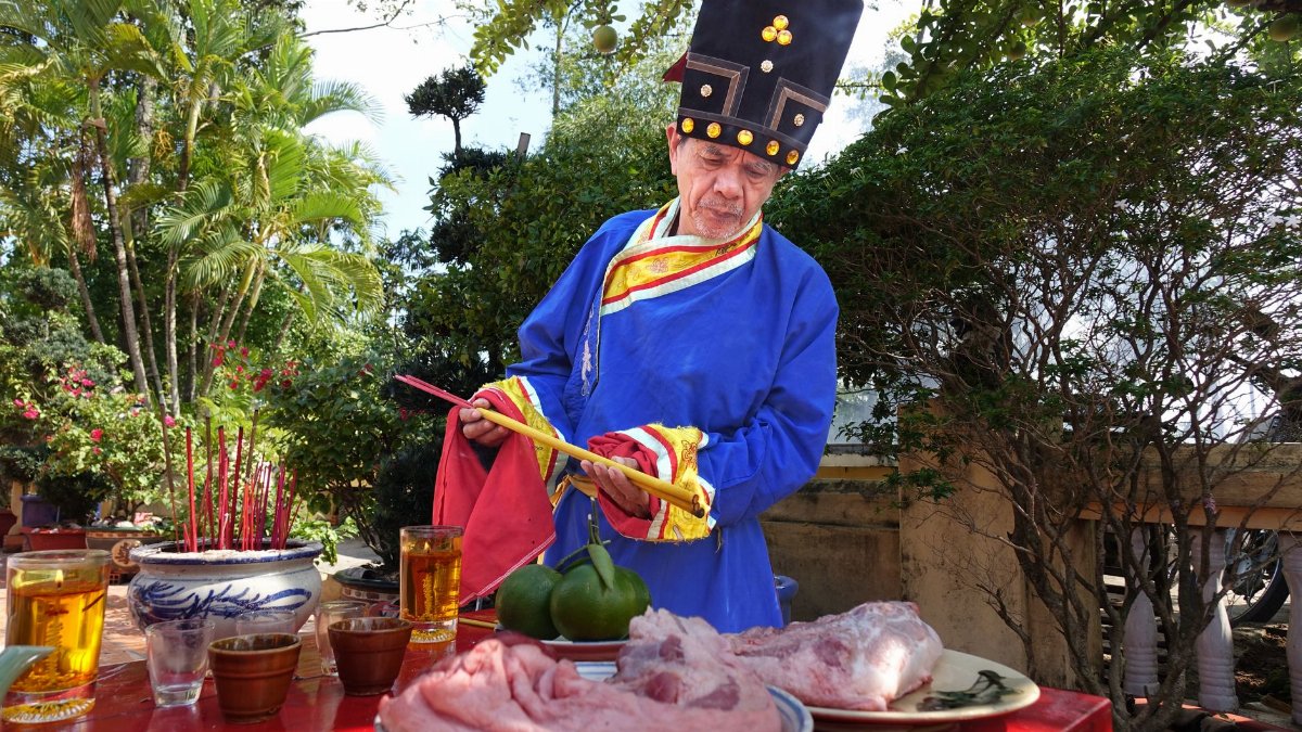 An Asian priest in traditional attire performs a ritual outdoors involving food offerings on a sunny day.
