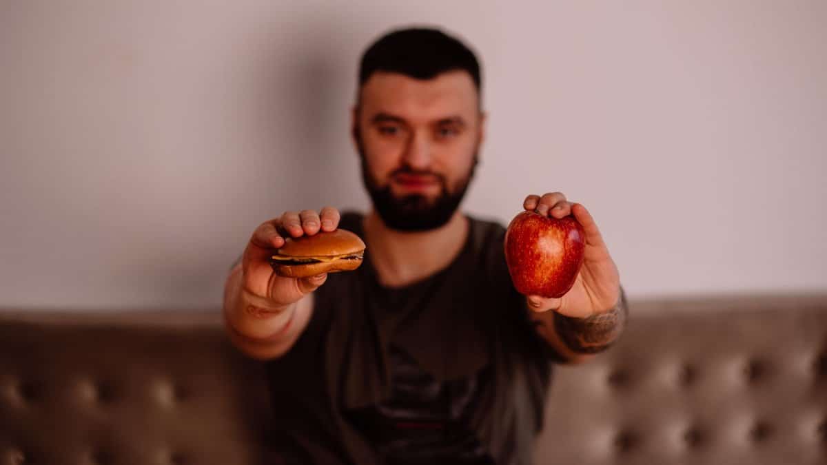 A man holds an apple and a cheeseburger, showcasing a choice between healthy and unhealthy food.