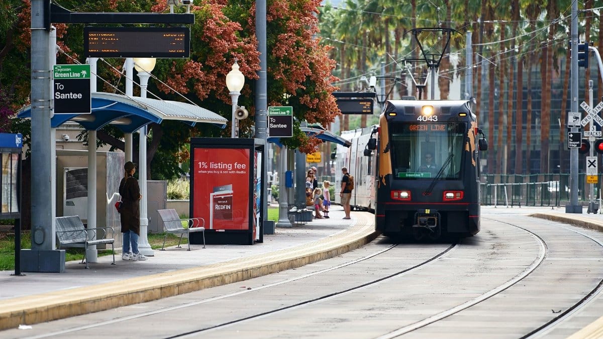 A modern tram arriving at a San Diego Green Line station with passengers waiting on a sunny day.