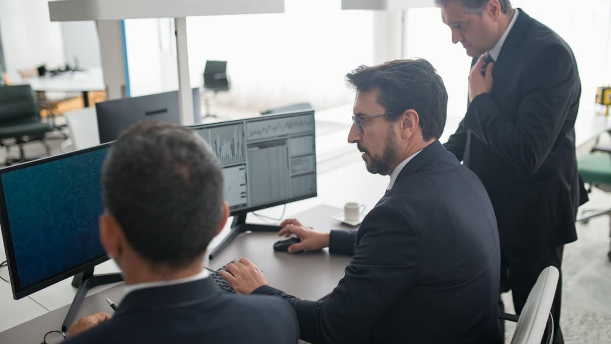 Colleagues in a modern office reviewing stock market trends and data on multiple screens.