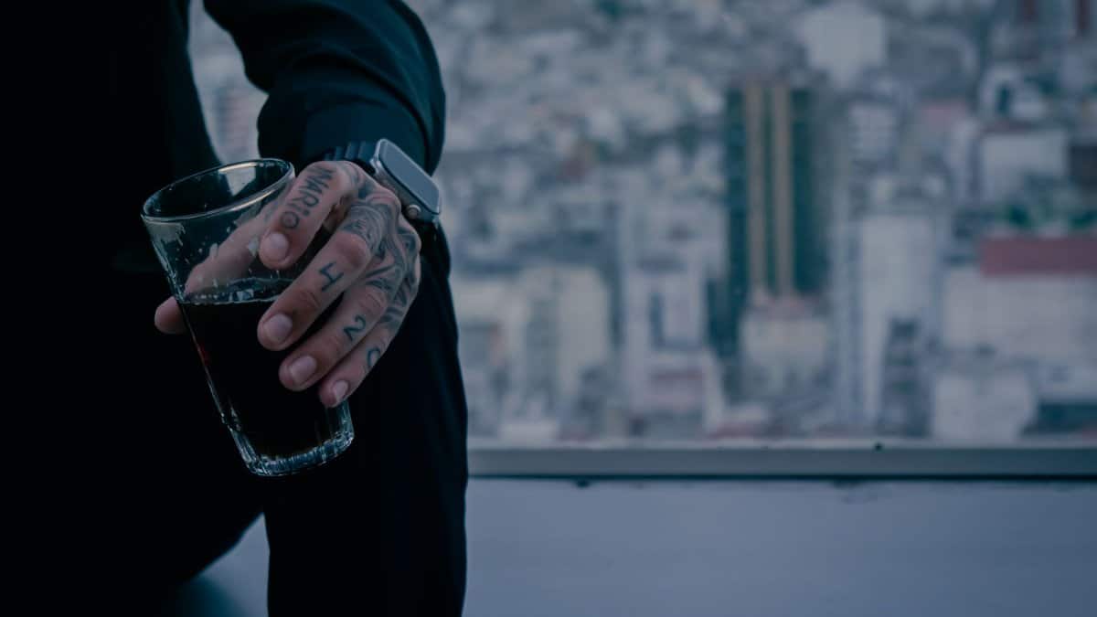 A tattooed hand holding a dark beverage in a glass with a blurred cityscape in the background.