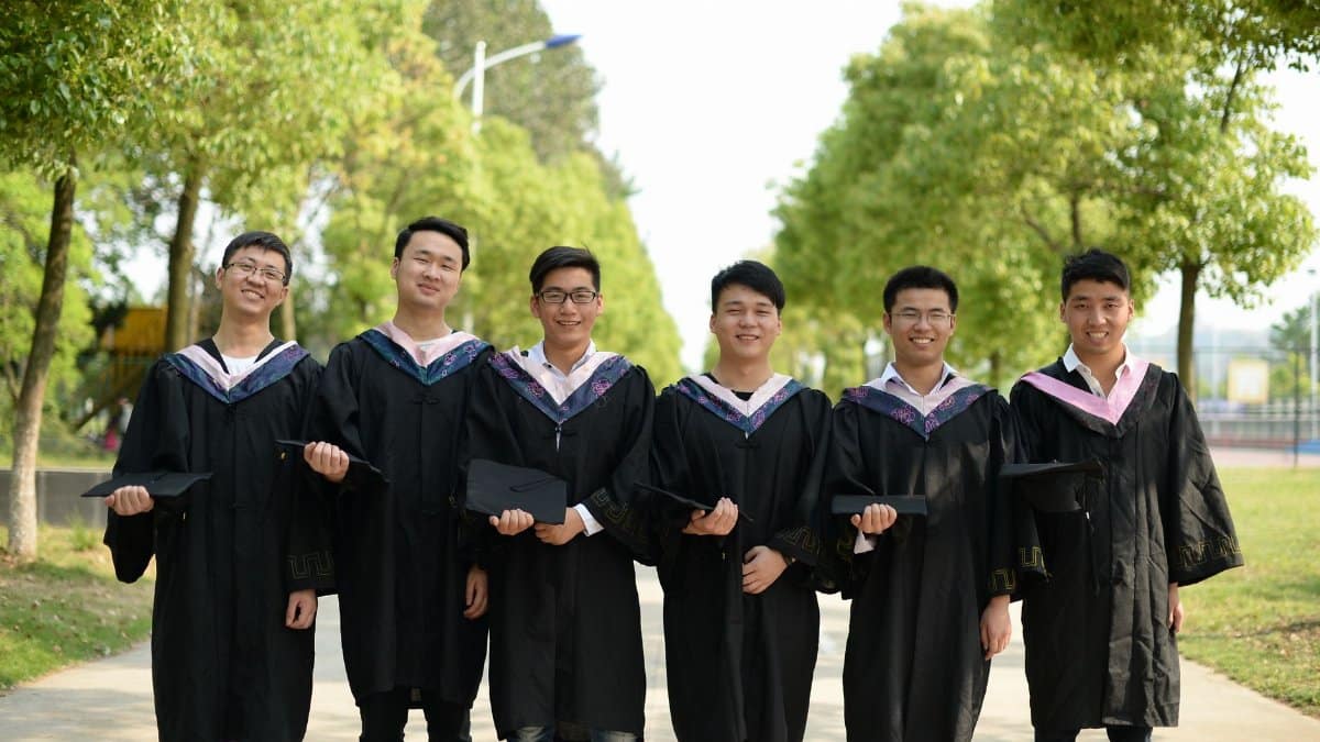 Six young graduates in gowns smiling and holding caps outdoors on a sunny day.