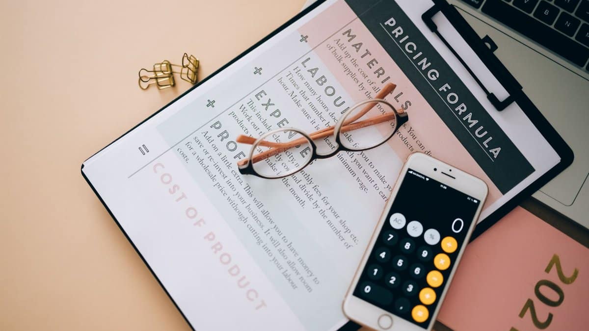 Overhead view of business tools including a phone calculator, pricing formula document, and eyeglasses on a desk.