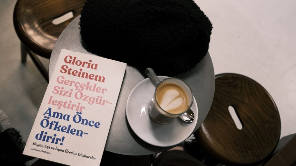A café setting with a latte, book by Gloria Steinem, and cozy hat on a table.