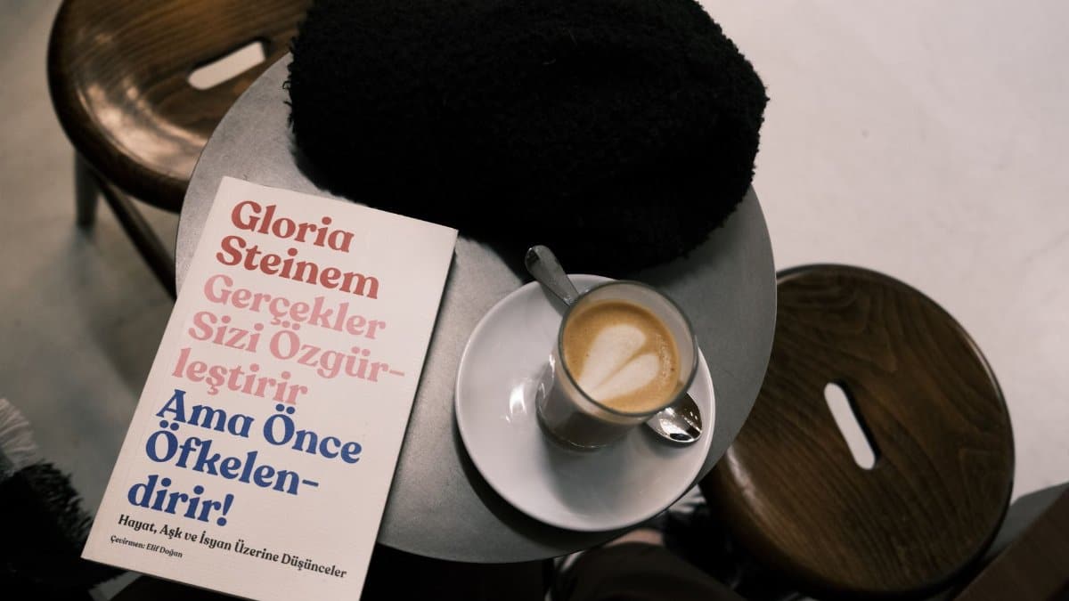 A café setting with a latte, book by Gloria Steinem, and cozy hat on a table.